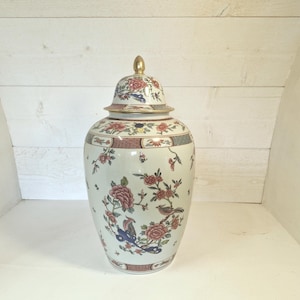 Puede incluir: Un tarro de jengibre de cerámica color crema con tapa a juego y remate dorado. El recipiente está decorado con un patrón colorido de peonías rosas, follaje verde y pájaros pequeños, acentuado por bordes decorativos en tonos rojos y azules.