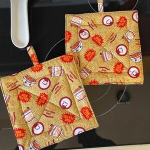 May include: Two yellow and red pot holders with a ramen noodle pattern. The pot holders have the words "Caution Hot" and "Rich Flavor" printed on them.