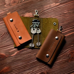 May include: Two brown leather keychains with the names "Linda" and "Brandon" embossed on them. The keychains are on a wooden surface and one keychain has a set of keys attached.