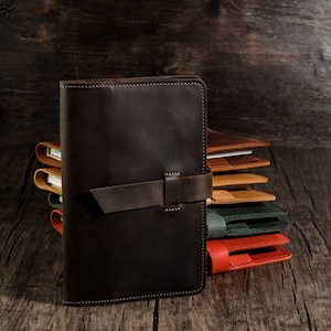 May include: A stack of leather journals in various colors, including brown, tan, green, and red. The front journal is dark brown with a strap closure. The journals are arranged on a wooden surface.