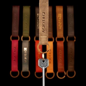 May include: Seven leather keyrings with gold rings and a silver key. The keyrings are in different colours: brown, green, red, orange, and black. The keyrings are all labelled with the word "AGIOTAGE".