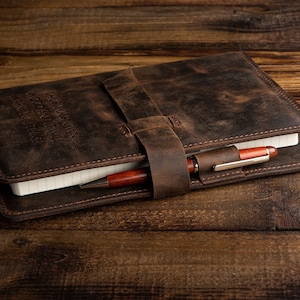 May include: A brown leather journal with a pen tucked into the strap. The journal has the words "Geography, History, Words, and Time, Learn to Trace" embossed on the cover.