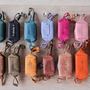 Könnte beinhalten: Eine Sammlung farbenfroher Leder-Kotbeutelhalter. Jeder Halter hat eine andere Farbe, darunter Türkis, Marineblau, Blau, Lavendel, Rosa und Rot. Die Halter sind mit Namen wie "Dolly", "Honey" und "Astra" personalisiert. Sie haben einen Metallverschluss und eine Schlaufe.