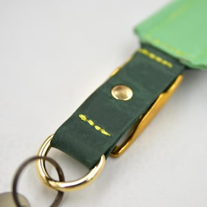 Puede incluir: Un llavero con una correa de cuero verde oscuro con costuras de color amarillo brillante y un remache de metal dorado. La correa est&aacute; unida a un anillo y un gancho de metal dorado pulido, con una secci&oacute;n de cuero verde claro visible arriba.