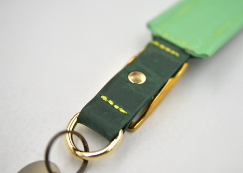 Puede incluir: Un llavero con una correa de cuero verde oscuro con costuras de color amarillo brillante y un remache de metal dorado. La correa est&aacute; unida a un anillo y un gancho de metal dorado pulido, con una secci&oacute;n de cuero verde claro visible arriba.