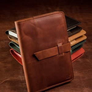 May include: A brown leather journal with a strap closure and the initials "KLF" embossed on the front. The journal is stacked on top of other journals in various colors.