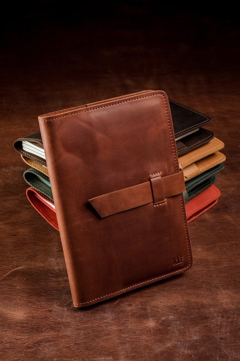 May include: A brown leather journal with a strap closure and the initials "KLF" embossed on the front. The journal is stacked on top of other journals in various colors.