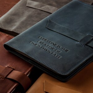 May include: A collection of leather journals in various colors, including blue, brown, and gray. The blue journal features the embossed text "EVERYONE IS THE CREATOR OF ONE'S OWN FATE." Each journal has a strap closure and stitched edges.