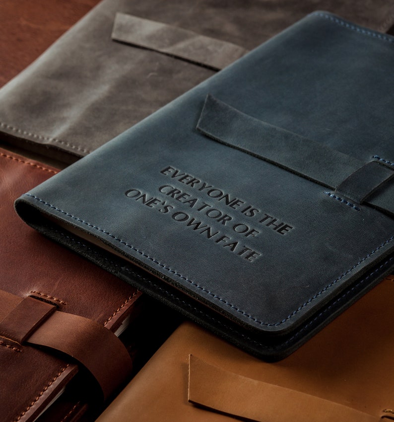 May include: A collection of leather journals in various colors, including blue, brown, and gray. The blue journal features the embossed text "EVERYONE IS THE CREATOR OF ONE'S OWN FATE." Each journal has a strap closure and stitched edges.