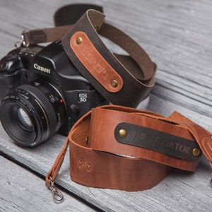 May include: Two brown leather camera straps with metal clasps. One strap has the text "JONOF1" engraved on it, and the other strap has the text "THE CREATOR" engraved on it.