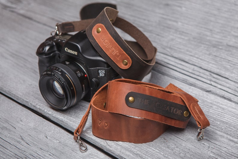 May include: Two brown leather camera straps with metal clasps. One strap has the text "JONOF1" engraved on it, and the other strap has the text "THE CREATOR" engraved on it.