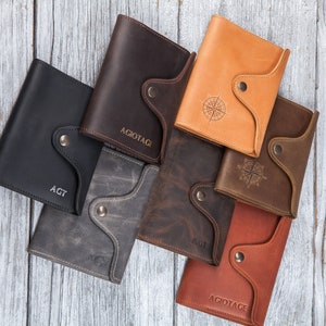 May include: A collection of leather wallets in various colors, including black, gray, brown, and tan. Some wallets feature engraved initials or a compass rose design. Each wallet has a button closure.