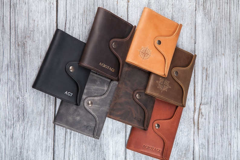 May include: A collection of leather wallets in various colors, including black, gray, brown, and tan. Some wallets feature engraved initials or a compass rose design. Each wallet has a button closure.