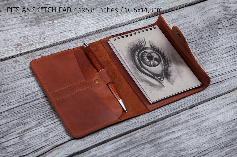 May include: Open brown leather sketch pad with a spiral-bound notepad featuring an eye drawing. A wooden pen is secured in a loop. The pad fits a 4.1 x 5.8 inch sketch pad. The text "FITS A6 SKETCH PAD 4,1x5,8 inches / 10,5x14,8cm" is visible.