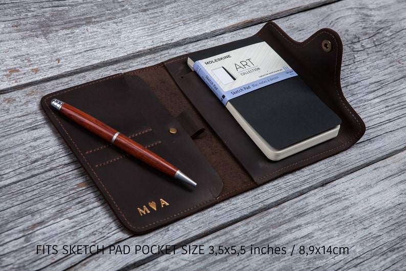 May include: A dark brown leather notebook cover with a matching pen and a black Moleskine sketchbook. The cover has a snap closure and a pocket for the pen. The pen is brown with silver accents. The notebook is 3.5 x 5.5 inches.