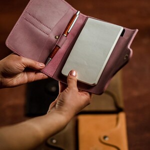 May include: A dusty rose leather notebook cover with a pen and a notepad. The cover is open, revealing the interior with a pen holder and a notepad. Other leather notebooks in different colors are visible in the background.