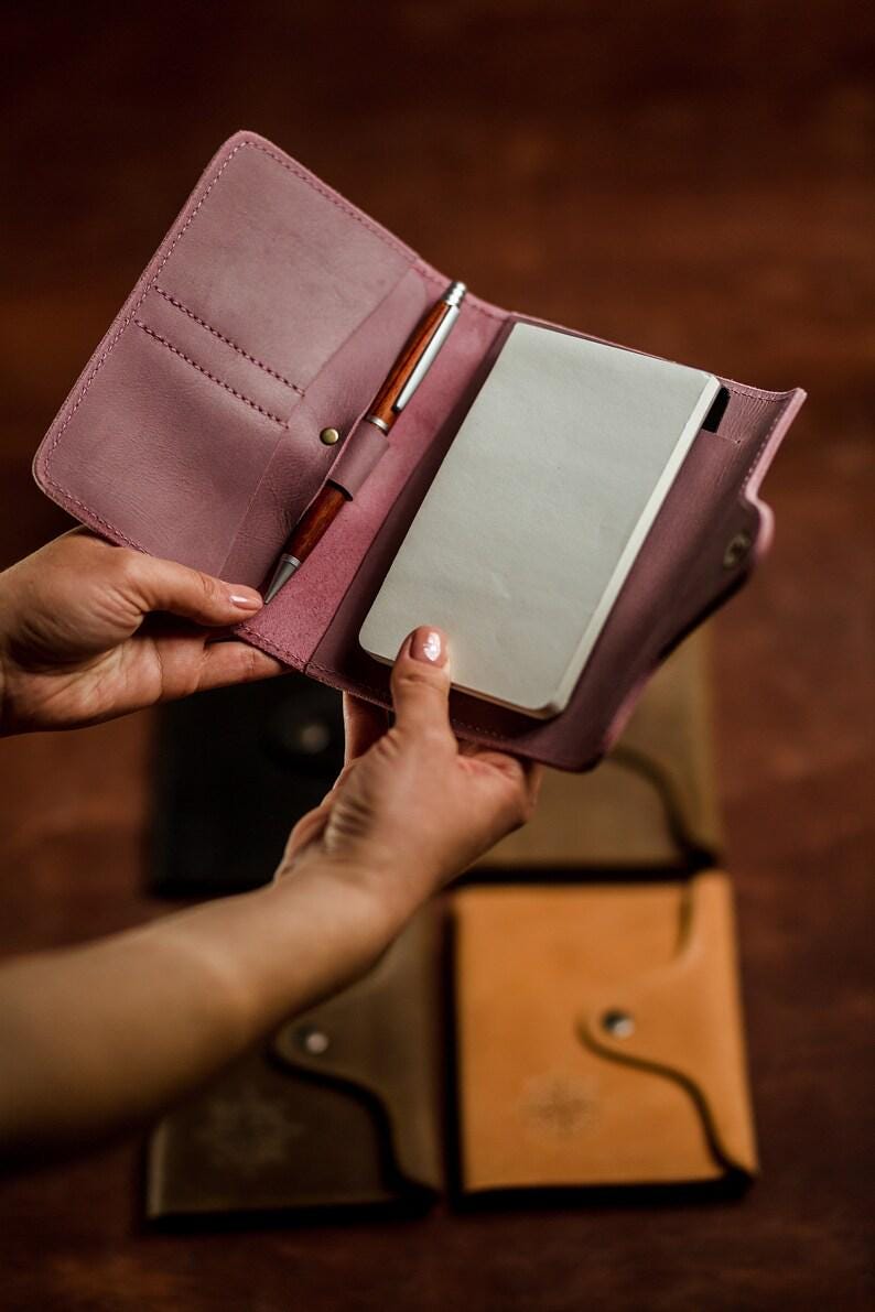May include: A dusty rose leather notebook cover with a pen and a notepad. The cover is open, revealing the interior with a pen holder and a notepad. Other leather notebooks in different colors are visible in the background.