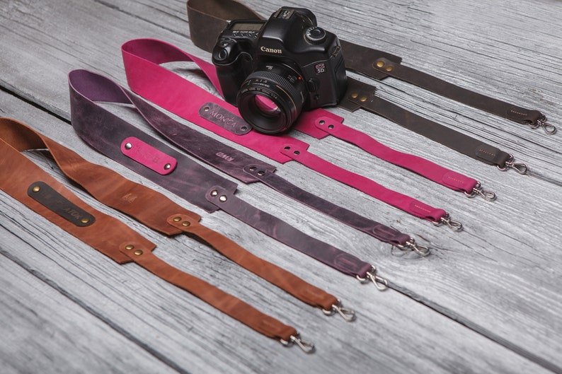 May include: Five leather camera straps in different colors, including brown, purple, and pink. Each strap has a metal clasp and a personalized name tag.