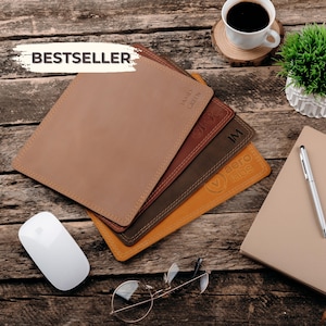 May include: A stack of four rectangular leather mouse pads in shades of tan, reddish-brown, dark brown, and mustard yellow, featuring stitched edges and personalised engravings including the text James Green, initials, and the logo aero labs.