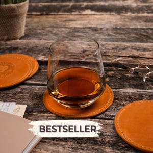 May include: A clear glass tumbler filled with amber liquid resting on a round tan leather coaster with visible stitching. The scene includes additional leather coasters, a pair of wire-rimmed glasses, and a white label with the text BESTSELLER.