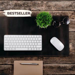 May include: A dark brown leather desk mat featuring the text BESTSELLER in the top left corner and M&R embossed in the bottom right. The mat holds a white wireless keyboard, a white wireless mouse, round-framed glasses, and a small green plant.