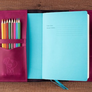 May include: A pink leather journal with a blue interior and a pen and colored pencils in the side pockets. The journal has a small embossed design of a person holding a heart.