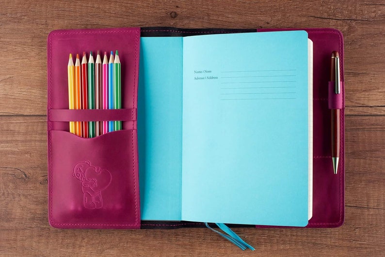 May include: A pink leather journal with a blue interior and a pen and colored pencils in the side pockets. The journal has a small embossed design of a person holding a heart.