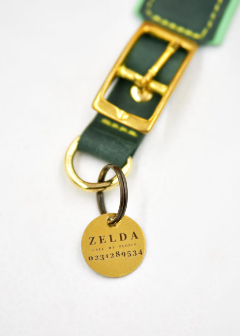 Puede incluir: Un collar de cuero verde oscuro con una hebilla de metal dorado y una anilla en D. Sujeta a la anilla en D hay una placa redonda de metal dorado grabada con el texto ZELDA, CALL MY PEOPLE y el n&uacute;mero de tel&eacute;fono 0231289534 en letras negras.