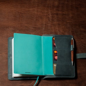 May include: A teal blue leather-bound journal with a pen holder and card slots. The journal is open to a blank page and has the text "LEUCHTTURM1917" printed on the bottom.