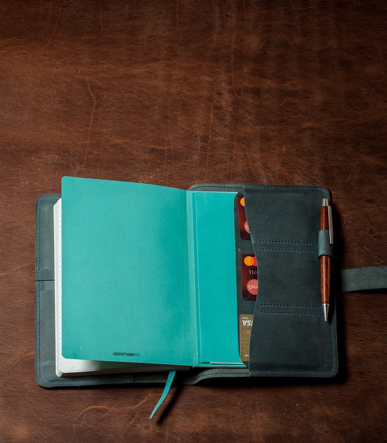 May include: A teal blue leather-bound journal with a pen holder and card slots. The journal is open to a blank page and has the text "LEUCHTTURM1917" printed on the bottom.