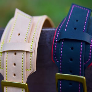 Op de afbeelding: Twee leren hondenhalsbanden, een is beige met gele stiksels en de andere is zwart met roze stiksels. Beide halsbanden hebben een gouden metalen gesp.