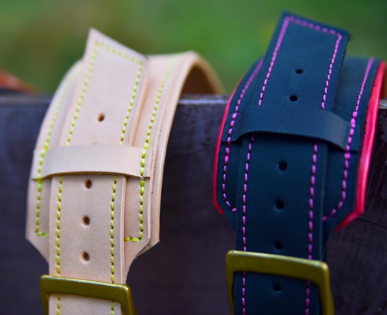 Op de afbeelding: Twee leren hondenhalsbanden, een is beige met gele stiksels en de andere is zwart met roze stiksels. Beide halsbanden hebben een gouden metalen gesp.