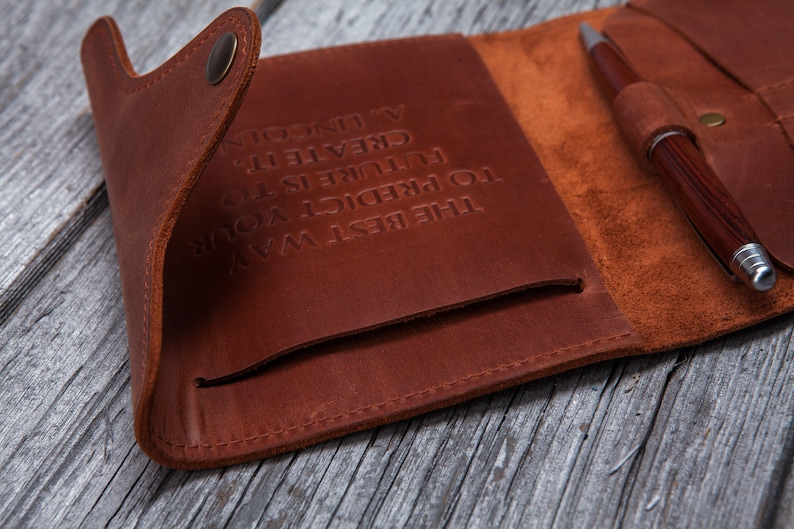 May include: A brown leather journal with a pen. The journal is open, revealing a pocket and the embossed quote: "THE BEST WAY TO PREDICT YOUR FUTURE IS TO CREATE IT. - A. LINCOLN." The pen is brown with silver accents.