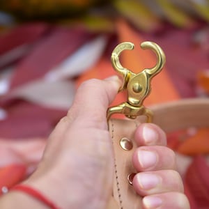 May include: A brass-colored swivel clasp with a curved design, attached to a light brown leather strap. The clasp is held against a blurred background of red and orange leaves, suggesting an autumn theme.