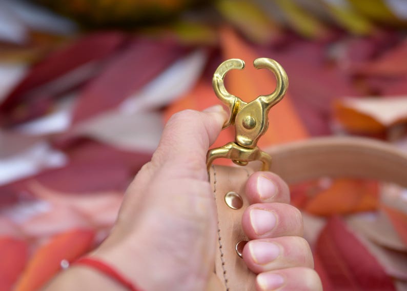 May include: A brass-colored swivel clasp with a curved design, attached to a light brown leather strap. The clasp is held against a blurred background of red and orange leaves, suggesting an autumn theme.