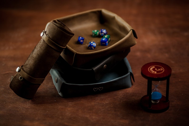May include: A set of three brown leather dice trays, one with blue and green dice, stacked on top of each other. A brown leather dice rolling tube is to the left of the trays. A small wooden hourglass with blue sand is to the right of the trays.