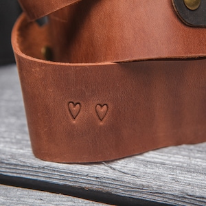 May include: A brown leather camera strap with two small heart-shaped cutouts.