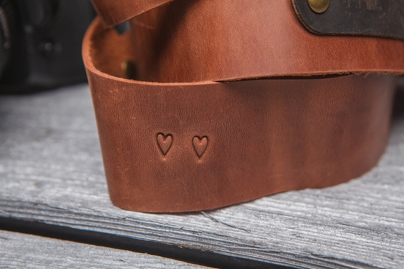 May include: A brown leather camera strap with two small heart-shaped cutouts.