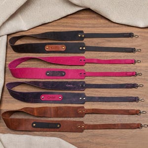 May include: Four leather camera straps in black, magenta, dark purple, and brown. Each strap features a central leather patch with a personalised name or initials, secured by two metal rivets on each side and metal swivel hooks at both ends.