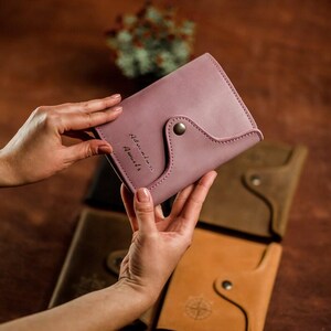 May include: A dusty rose leather wallet with a curved flap and snap closure. The text "Adventure Awaits" is stitched on the front. Other leather wallets in brown and tan are visible in the background.