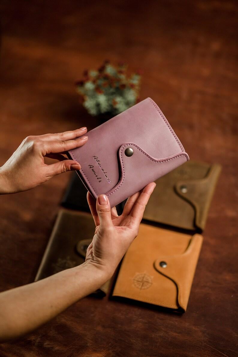 May include: A dusty rose leather wallet with a curved flap and snap closure. The text "Adventure Awaits" is stitched on the front. Other leather wallets in brown and tan are visible in the background.