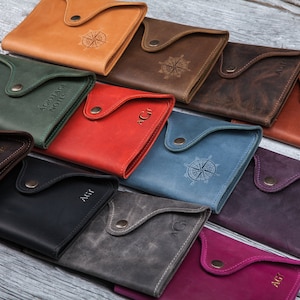 May include: A collection of leather wallets in various colors, including tan, green, red, and blue. Each wallet features a flap closure with a snap and a debossed design or initials. The wallets are arranged on a light wood surface.