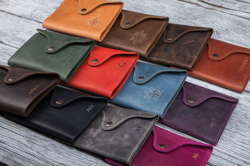 May include: A collection of leather wallets in various colors, including tan, green, red, and blue. Each wallet features a flap closure with a snap and a debossed design or initials. The wallets are arranged on a light wood surface.