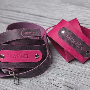 May include: Two personalized leather camera straps in shades of pink and purple. The strap on the left has the name "Rachel" embossed on it, while the strap on the right has the name "Monica" embossed on it.