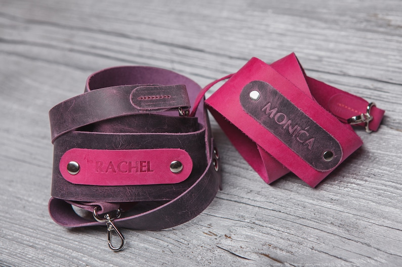 May include: Two personalized leather camera straps in shades of pink and purple. The strap on the left has the name "Rachel" embossed on it, while the strap on the right has the name "Monica" embossed on it.