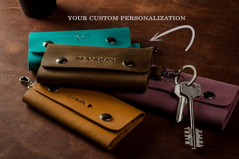 May include: Three leather keychains with different colors and personalized names. The keychain on top is teal blue with a monogram, the middle keychain is brown with the name "BRANDON" and the bottom keychain is tan with the name "Linda" and a heart. The keychains are attached to a silver key ring with two keys.