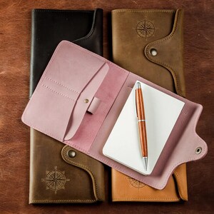 May include: A collection of leather notebooks in various colors, including black, brown, and pink. One notebook is open, revealing a blank notepad and a wooden pen. The notebooks have a compass design and are arranged on a brown leather surface.