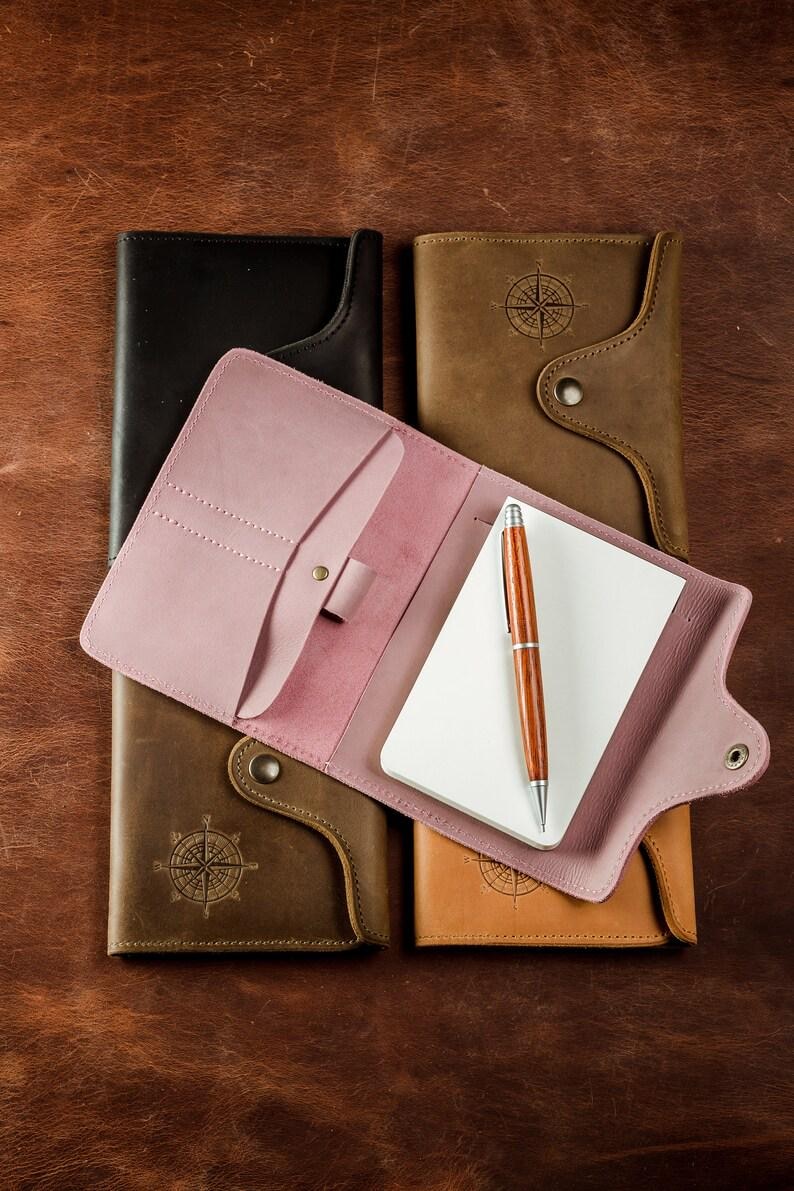 May include: A collection of leather notebooks in various colors, including black, brown, and pink. One notebook is open, revealing a blank notepad and a wooden pen. The notebooks have a compass design and are arranged on a brown leather surface.
