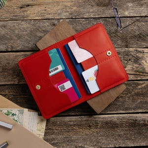May include: A red leather wallet, open to display several credit cards and identification cards. The wallet has a snap closure and is set on a wooden surface. The cards are various colors, including blue, green, and pink.