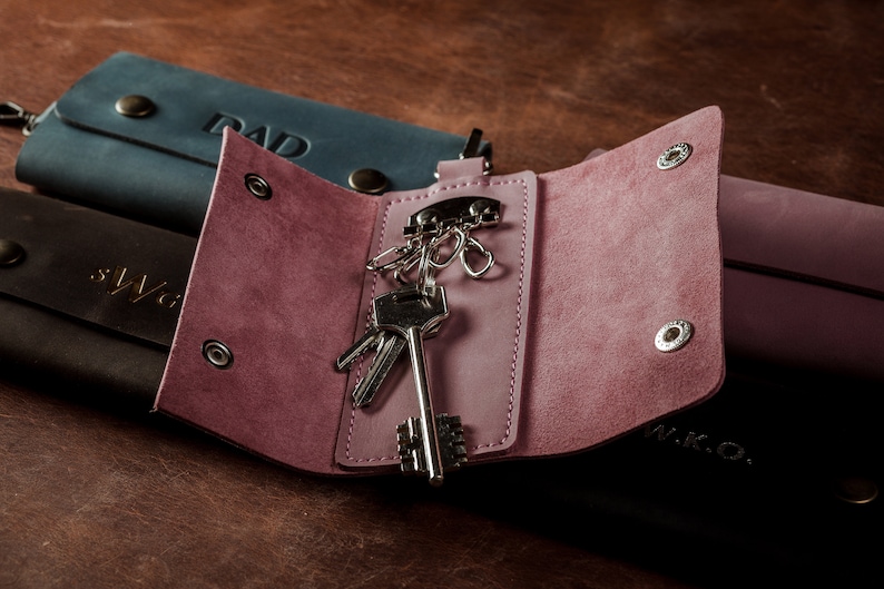 May include: A pink leather key holder with a silver key ring and keys inside. The key holder is open and the inside is visible. The key holder has two silver snaps on the outside.